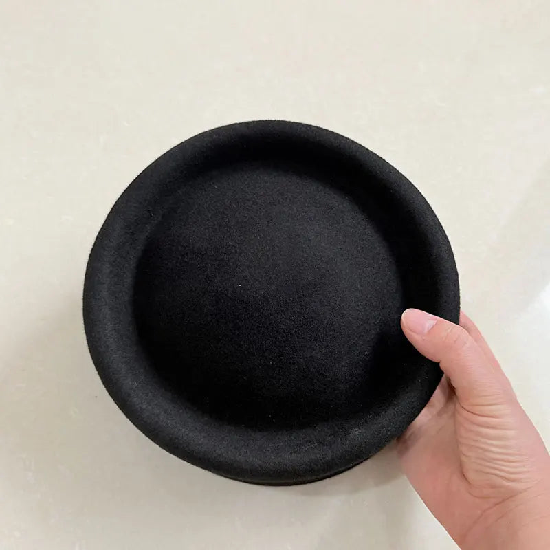 Black pillbox hat held by a hand on a light beige background