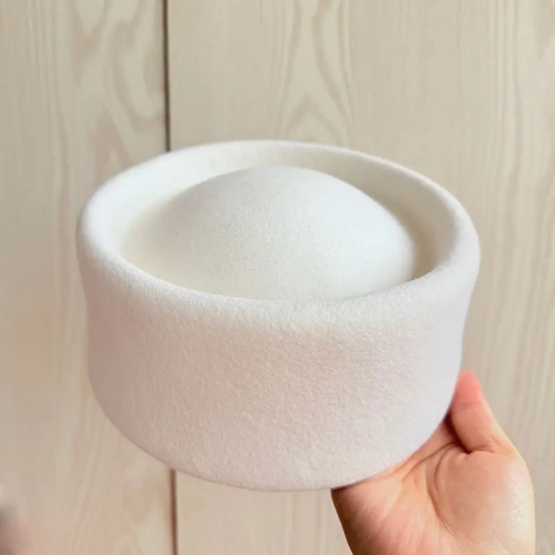 White pillbox hat held by a hand against a light wooden background